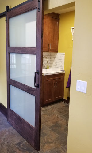 Photo peeking into laundry room with sink and white tile backsplash behind sliding barn door with frosted glass