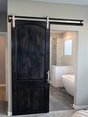 photo of a dark wood barn door opening up to a bathroom with a freestanding tub and shower with white tile half wall and frameless shower door