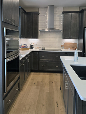 photo of kitchen with wood floors, dark cabinets, a stainless cooktop hood vent, and an island sink