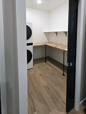 photo of laundry room with stacked washer and dryer, shelves and a wood countertop supported by metal legs, with wood floor