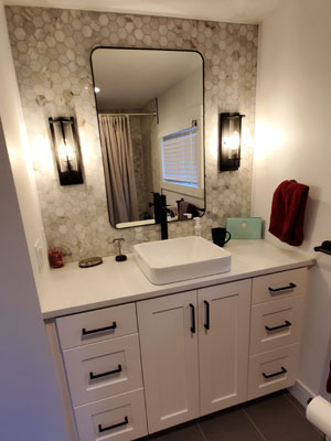 photo of bathroom with raised rectangle ceramic sink, stone honeycomb tile walls, black sconces, black fram rounded mirror and white cabinets