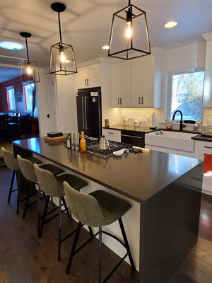 photo of kitchen island with black quartz waterfall countertop, green stools, farmhouse sink and black frame pendants